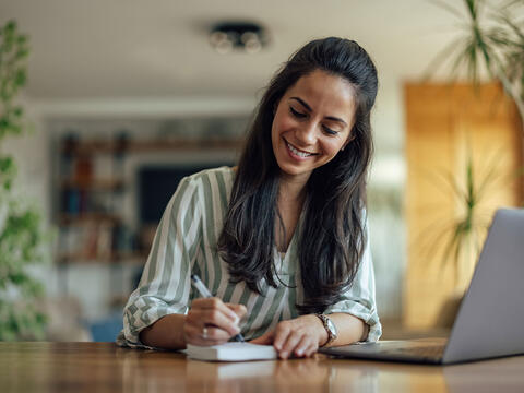 person sidder ved computer i sit hjem og noterer på papir
