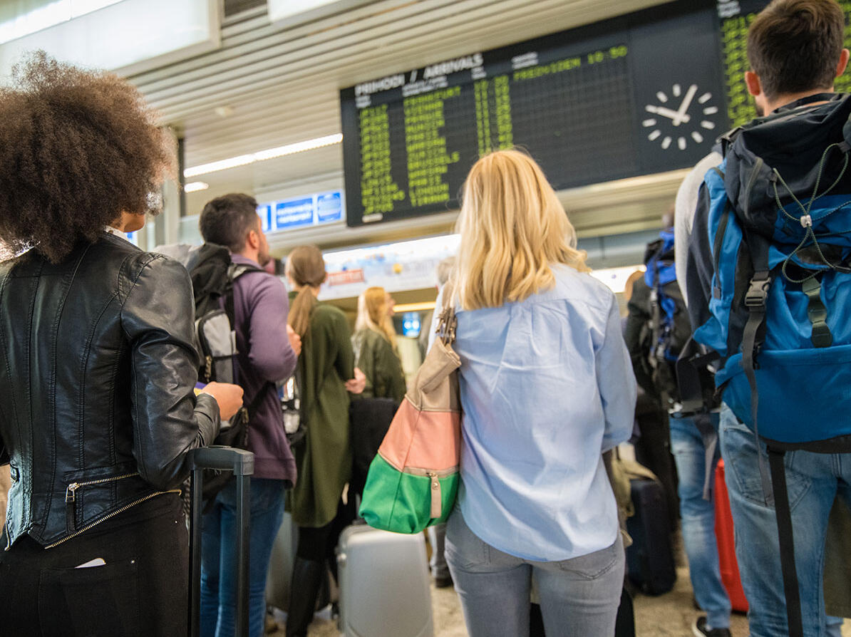 Personer står med deres bagage og kigger på en tavle