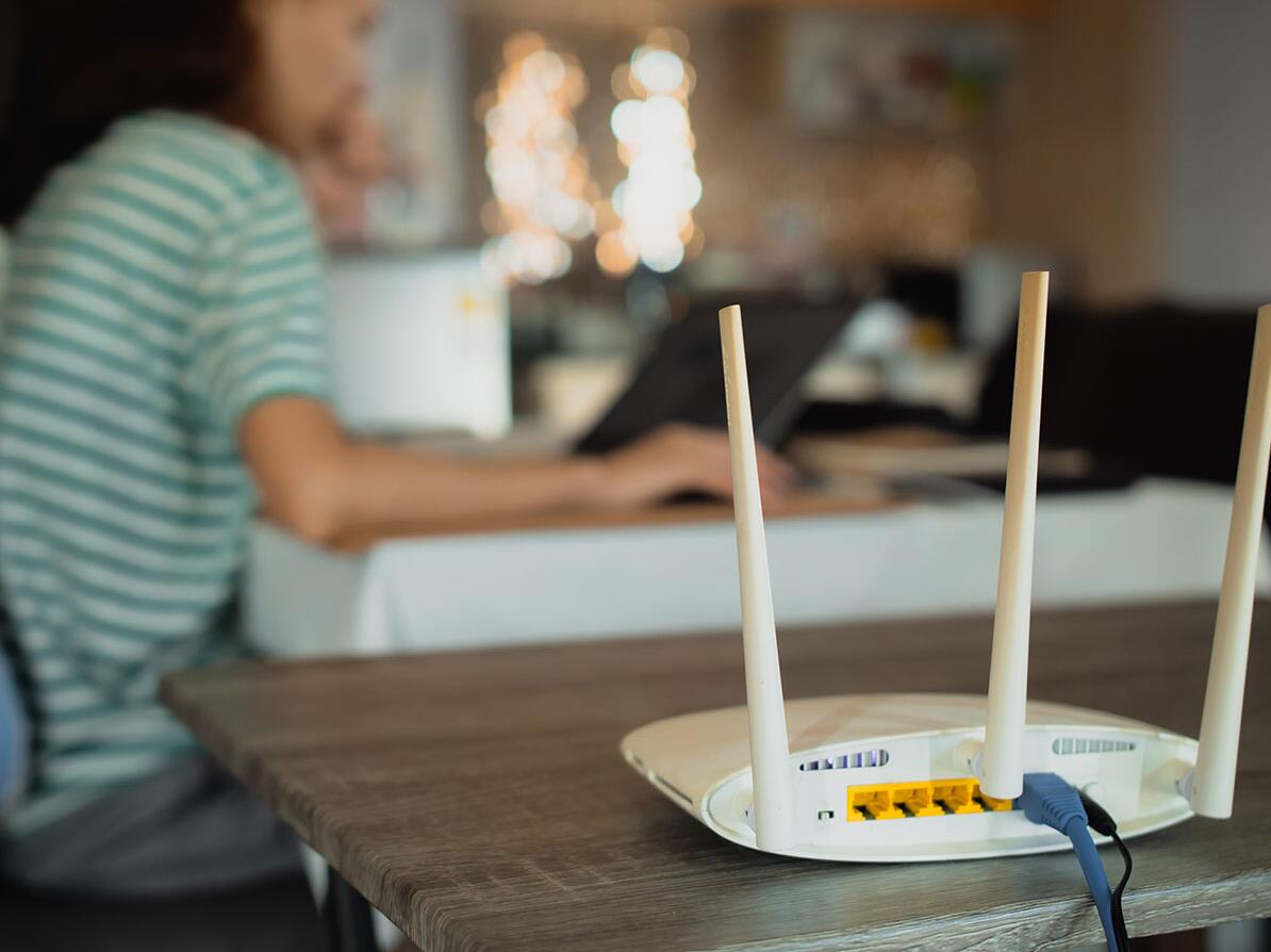 En router står på et bord, og en personer sidder med sin computer i baggrunden