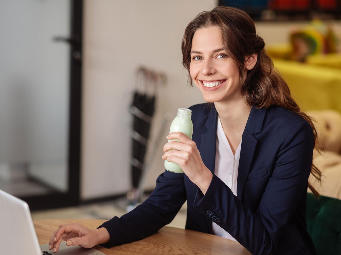kvinde sidder ved computer og drikker drikkeyoghurt.