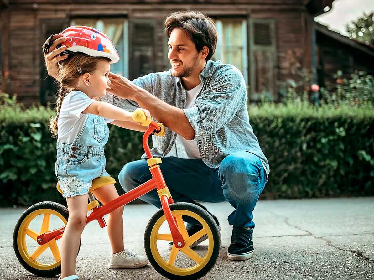 Pige sidder på løbecykel mens far giver hende cykelhjelm på