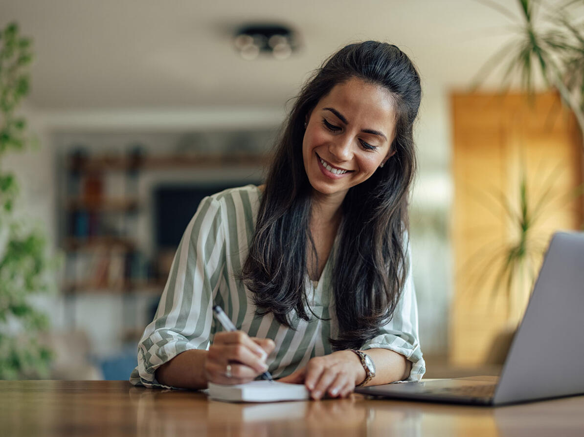 person sidder ved computer i sit hjem og noterer på papir