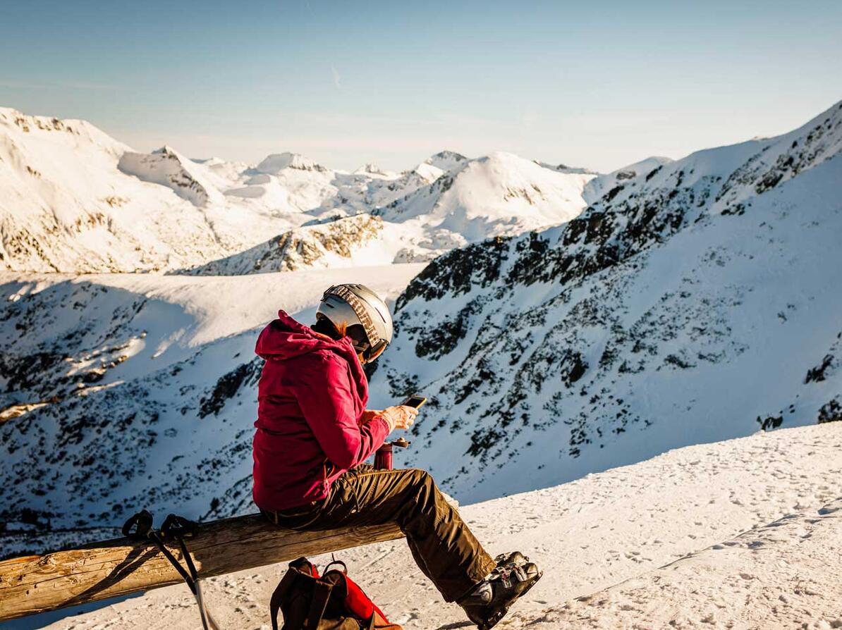 Person sidder i skitøj på sneklædt bjerg og kigger på sin telefon