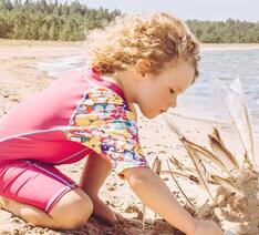Barn med soltøj sidder i sand ved havet.