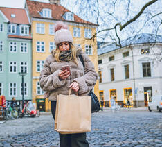 Person i vintertøj står med papirspose i den ene hånd og smartphone i den anden hånd, i København