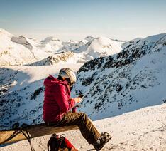 Person sidder i skitøj på sneklædt bjerg og kigger på sin telefon